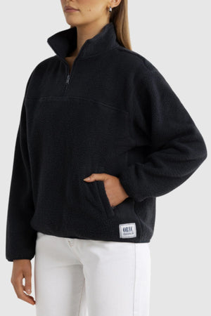 Someone wearing the ORTC Sherpa Fleece Quarter Zip in black, featuring a front pocket and a small rectangular patch near the hem, paired with white pants against a plain light gray background.