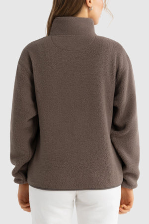 A person is seen from the back wearing the ORTC Sherpa Fleece Quarter Zip in brown with a high collar and long sleeves, paired with white pants against a plain light gray background.