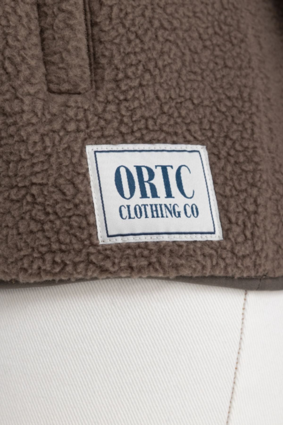 A close-up of the ORTC Sherpa Fleece Quarter Zip in brown, featuring a white rectangular “ORTC CLOTHING CO” patch in blue text, styled above a pair of white pants.