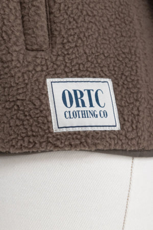 A close-up of the ORTC Sherpa Fleece Quarter Zip in brown, featuring a white rectangular “ORTC CLOTHING CO” patch in blue text, styled above a pair of white pants.