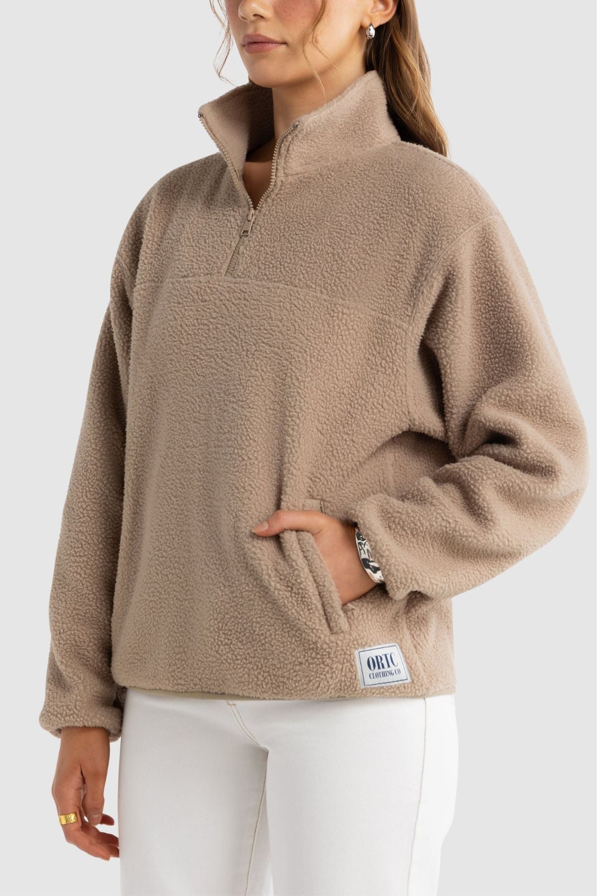 A woman with long brown hair wears the ORTC Sherpa Fleece Quarter Zip—a light brown pullover with a front pocket—paired with white pants, standing against a plain, light background.