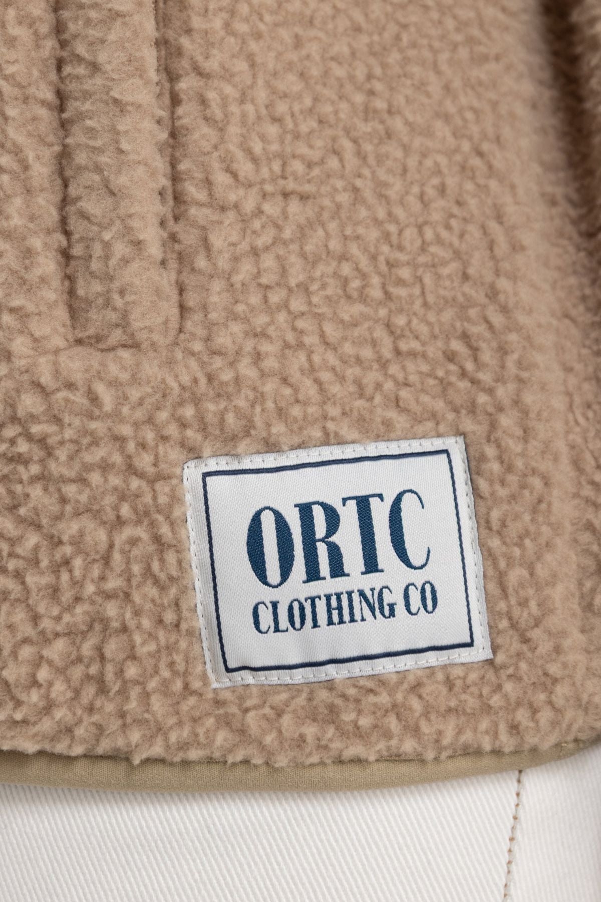 A close-up of the ORTC Sherpa Fleece Quarter Zip in beige, featuring a stitched white rectangular "ORTC CLOTHING CO" label near the bottom hem, paired with white pants.
