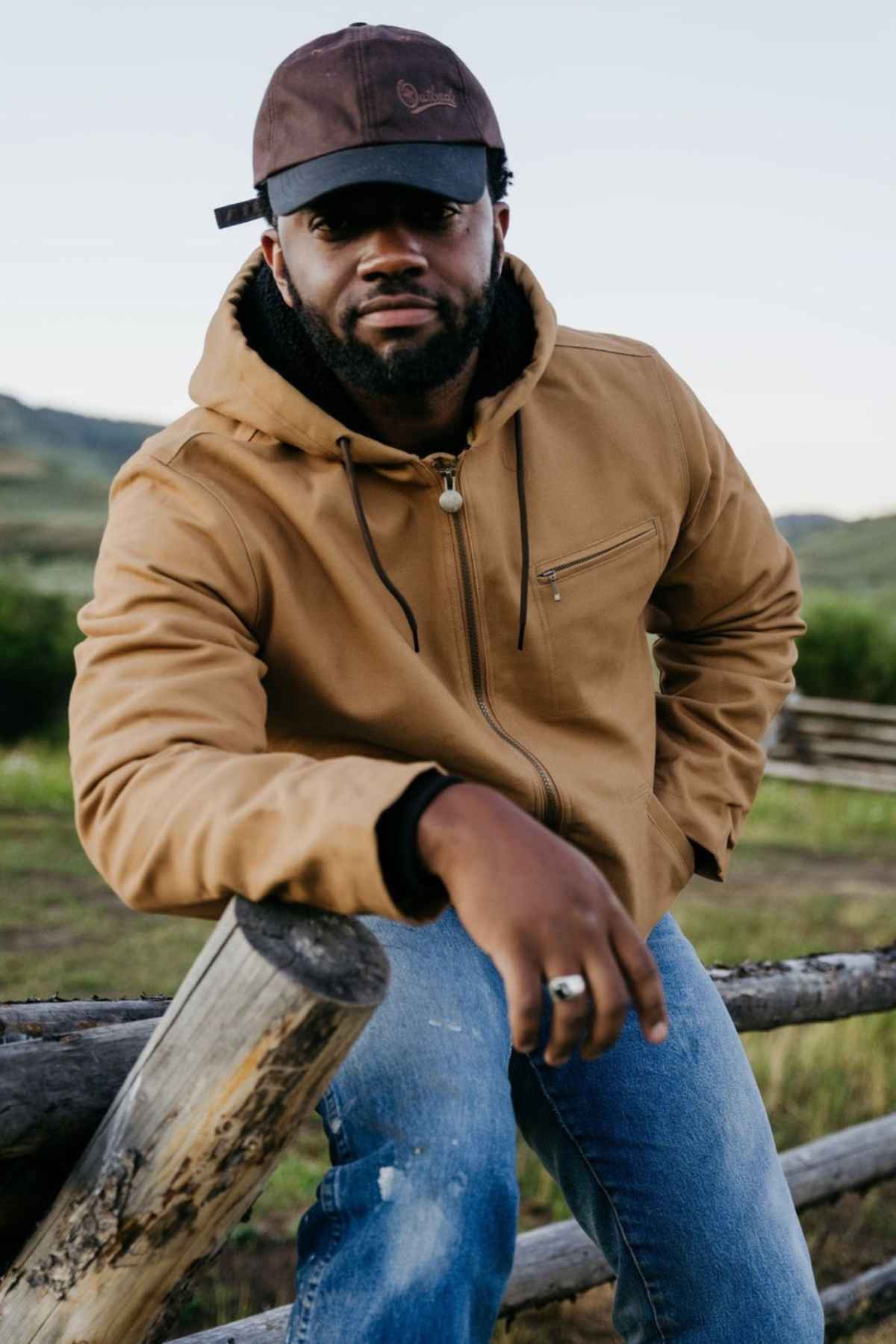 Wearing the Outback Canvas Sawbuck Hoodie, a man leans on a wooden fence against green hills and a cloudy sky, highlighting the hoodie’s rugged durability and water-resistant canvas fabric.