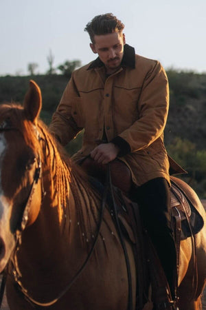 A man in an Outback Cattleman Jacket rides a brown horse outdoors, sunlight highlighting them with shrubs and a hill in the background.