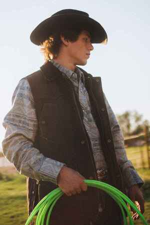 A person dressed in Western attire, including a cowboy hat, patterned shirt, and the Outback Cattleman Vest, stands outdoors holding a neon green lasso. The person is looking off to the side with a serious expression. In the background, there is grassy terrain and a fence.