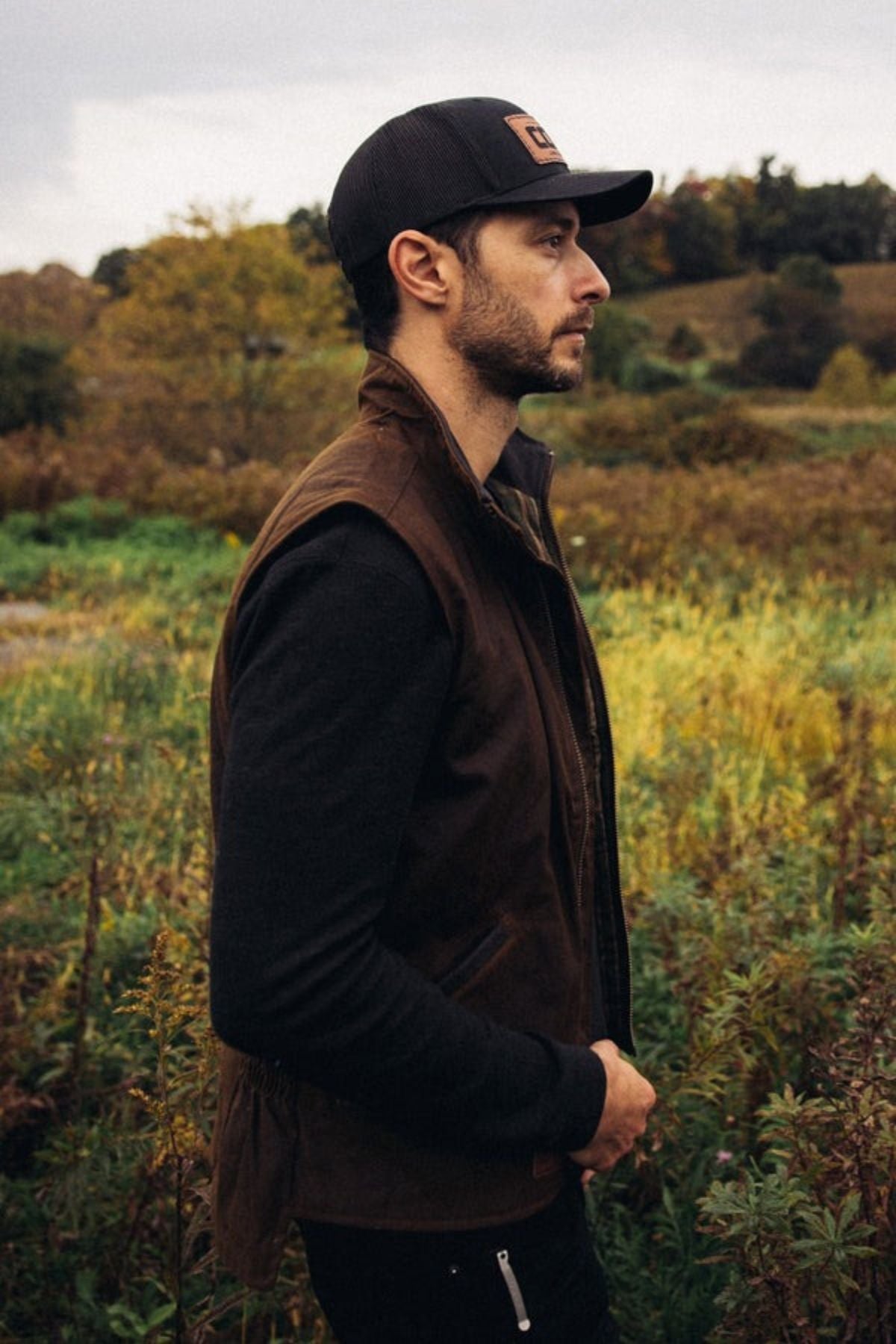 A man in a black cap, dark long-sleeve shirt, and the Outback Sawbuck Canyonland Vestβcrafted from brown oilskin for waterproof protectionβstands in profile in a grassy field with trees and cloudy skies, ready for outdoor adventures.