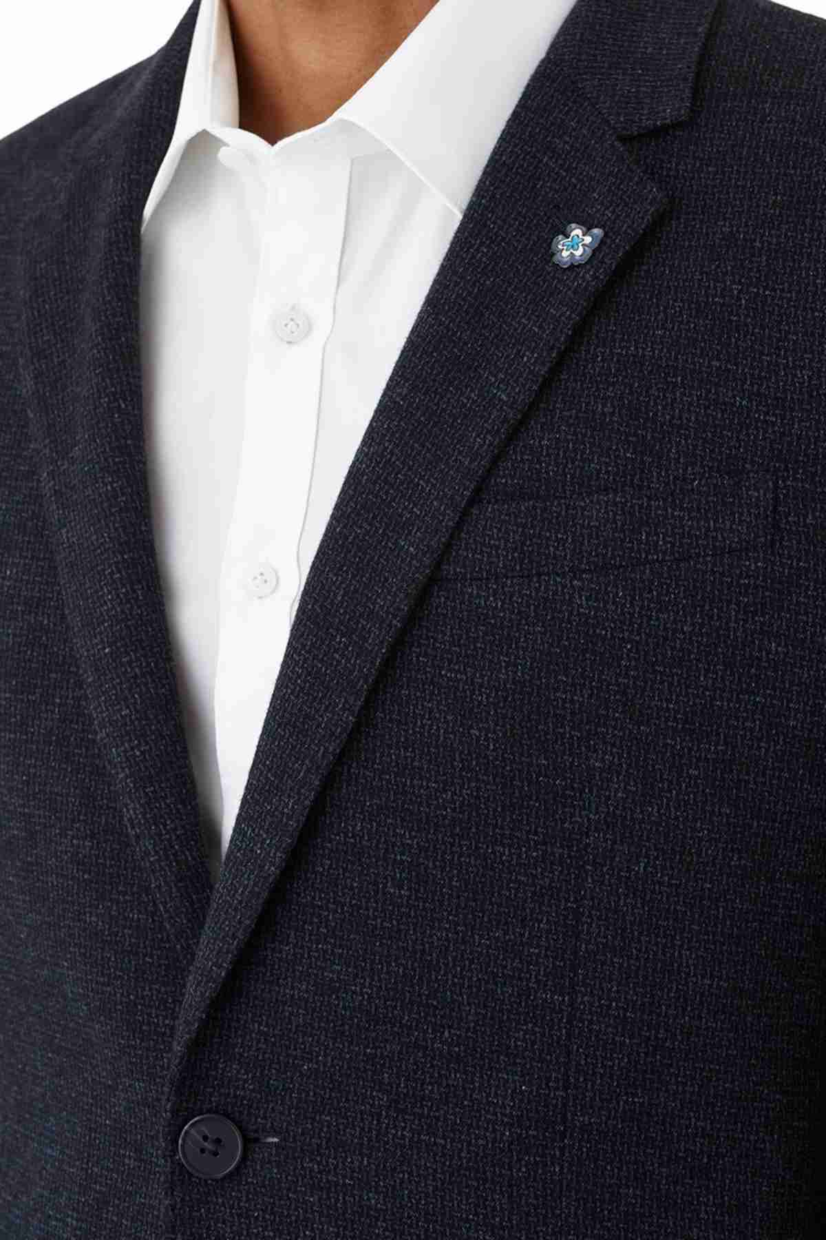 A close-up reveals a person wearing the Uberstone Zeller Jacket, a dark textured wool-blend blazer in a skinny fit. The jacket features visible collar and lapel details and is styled with one button fastened. It also showcases a small blue and silver floral lapel pin, adding elegance.