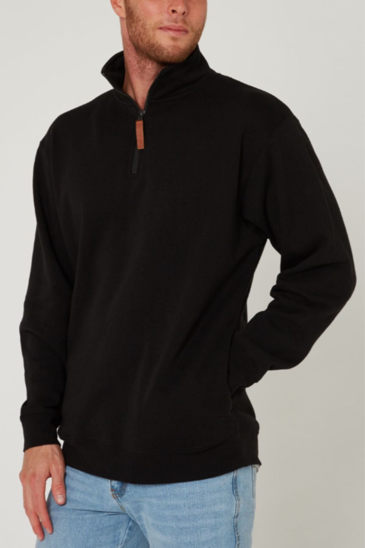 A man stands against a plain light-colored background, wearing the Pilbara Classic Fleece Pullover in black with cozy fleece lining and light blue jeans, one hand in his pocket.