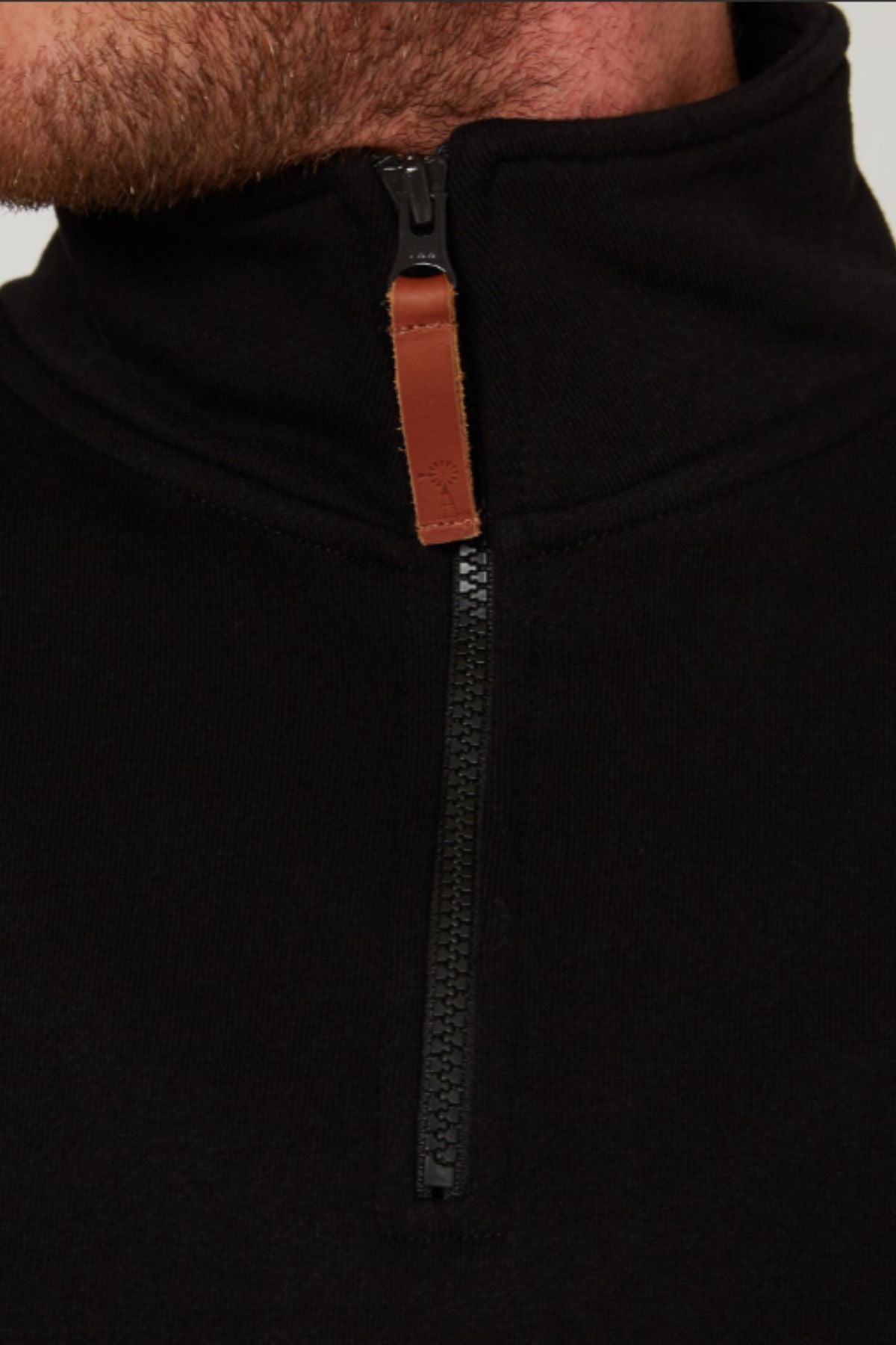 Close-up of a person wearing a black Pilbara Classic Fleece Pullover with a brown leather zipper pull tab. The image shows only the lower face, neck, and upper chest. Features plush fleece lining for extra warmth.