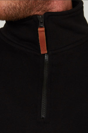 Close-up of a person wearing a black Pilbara Classic Fleece Pullover with a brown leather zipper pull tab. The image shows only the lower face, neck, and upper chest. Features plush fleece lining for extra warmth.