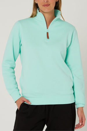 A woman wears the Pilbara Ladies Classic Fleece Pullover in light turquoise with cozy fleece lining and black pants, standing against a plain background with one hand in her pocket.