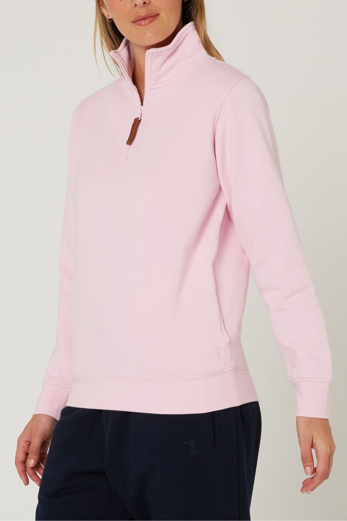A person wearing a light pink Pilbara Ladies Classic Fleece Pullover with a high collar and cozy fleece lining, paired with navy blue pants, stands against a plain, light-colored background.