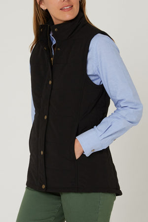 A person wears the Pilbara Ladies Vest, a black sleeveless microfibre waterproof winter vest with Polyfill padding, over a light blue button-up shirt and green pants, standing with one hand in their pocket against a plain background.