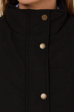 Close-up of a person wearing the Pilbara Ladies Vest, a black quilted vest with Polyfill padding, high collar, and gold snap buttons. Only the lower face and upper chest are shown.