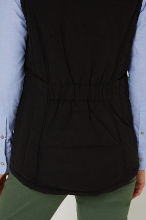 A person viewed from the back wears a light blue long-sleeve shirt, green pants, and the Pilbara Ladies Vest—a black winter vest with polyfill padding and an elastic waistband.