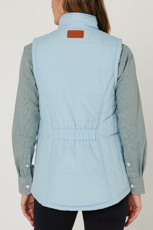 A person is shown from the back wearing the Pilbara Ladies Vest in light blue with Polyfill padding over a greenish-gray long-sleeve shirt. The vest has a high collar, fitted waist, and brown patch on the upper back.