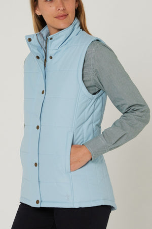 A woman wearing the Pilbara Ladies Vest in light blue with Polyfill padding over a gray-green long-sleeve shirt, one hand in her vest pocket, stands against a plain background.