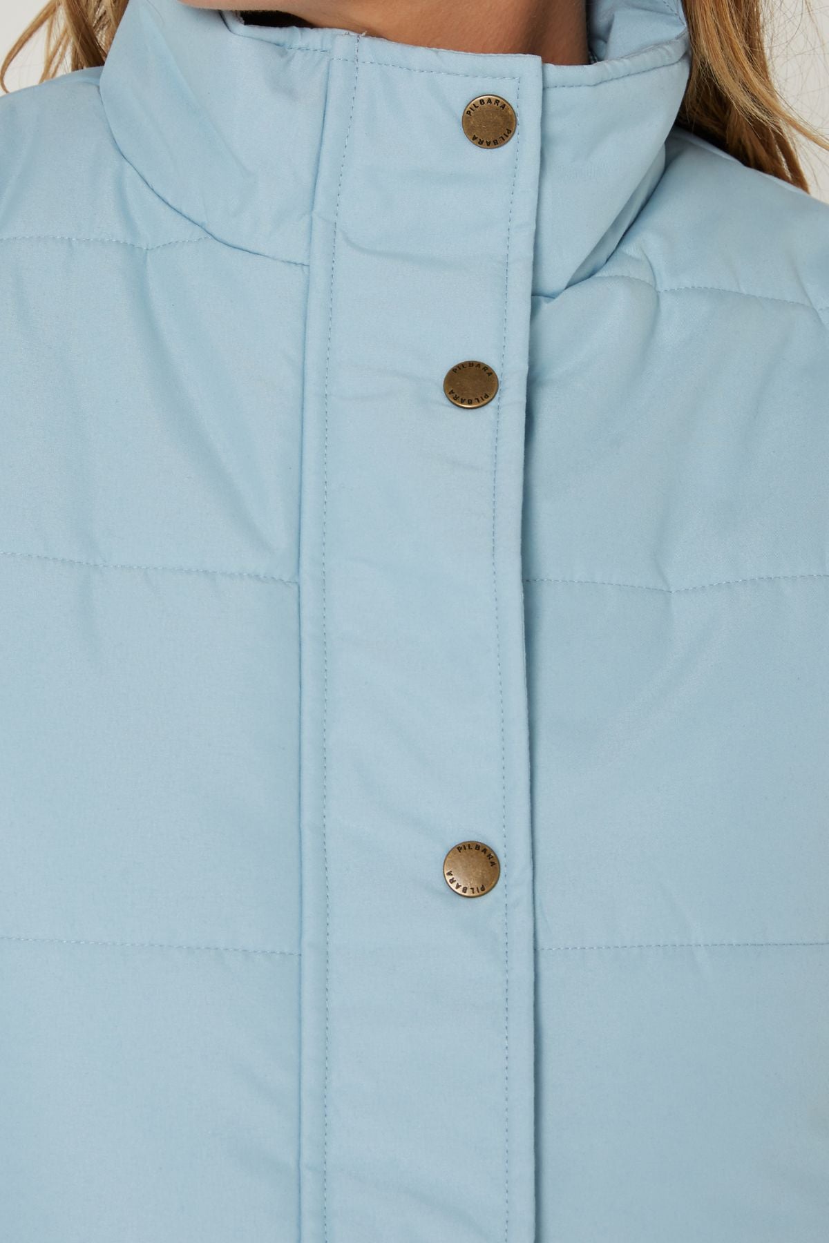 Close-up of a Pilbara Ladies Vest in light blue, featuring a quilted design, high collar, and metallic snap buttons. The image highlights the upper chest area along with part of the model's neck and hair.