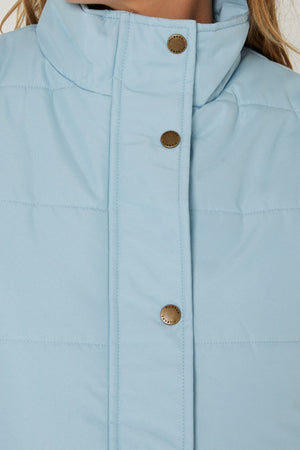 Close-up of a Pilbara Ladies Vest in light blue, featuring a quilted design, high collar, and metallic snap buttons. The image highlights the upper chest area along with part of the model's neck and hair.