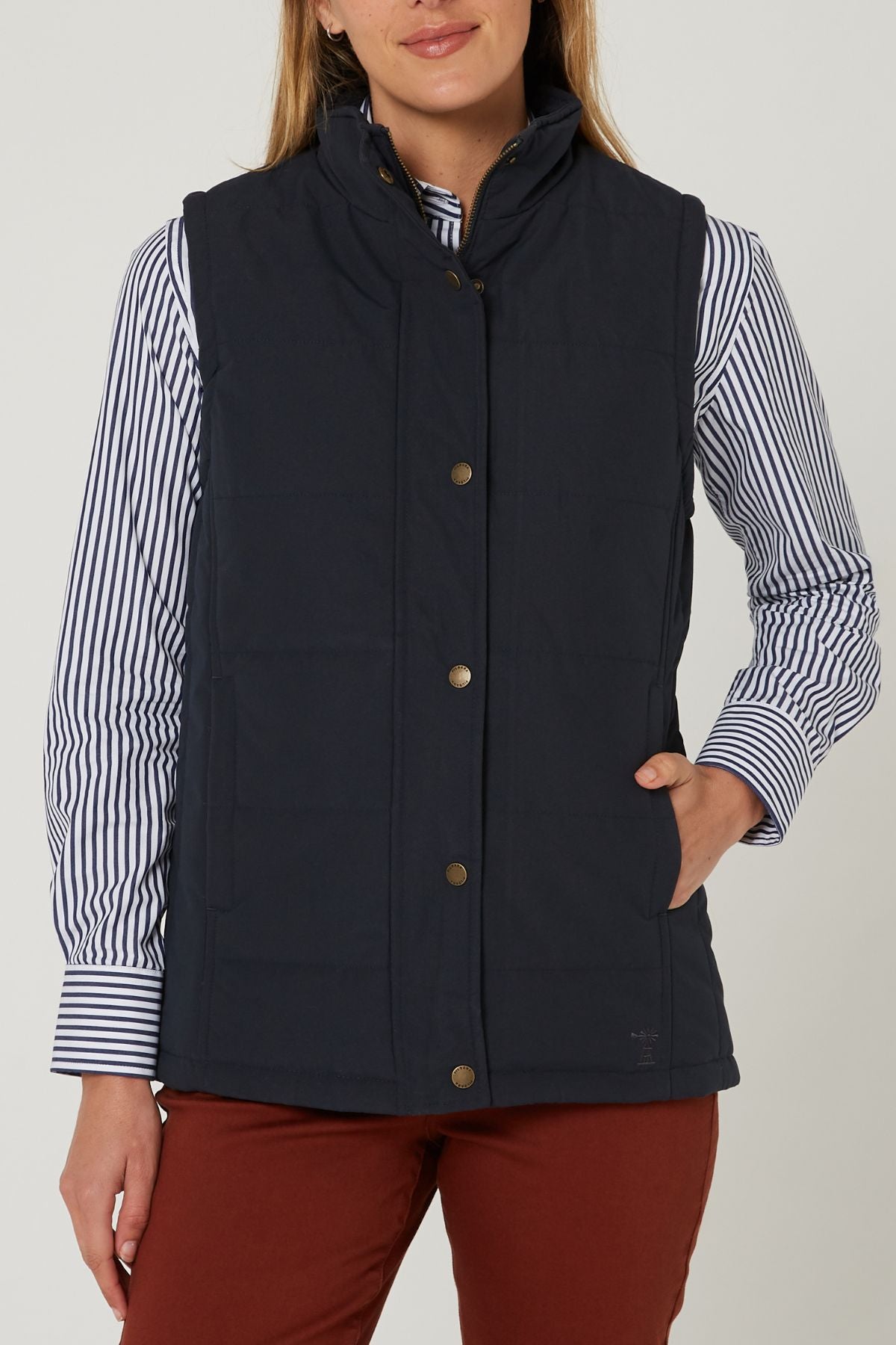 A woman wears the Pilbara Ladies Vest, a navy microfibre waterproof winter vest with polyfill padding, over a striped button-down shirt and rust-colored pants, standing with one hand in her pocket against a plain light background.