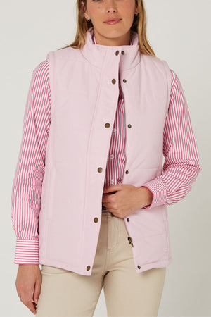 A woman stands against a plain background wearing a light pink Pilbara Ladies Vest over a pink and white striped shirt and beige pants, holding the vest near the zipper with her right hand.