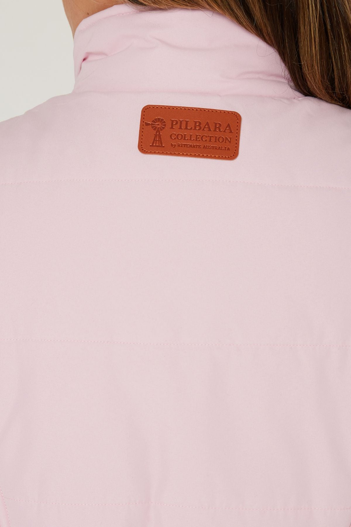 Close-up of the back of a light pink Pilbara Ladies Vest with Polyfill padding and a brown leather patch reading "Pilbara Collection by Statesman Australia." Part of a person’s neck and hair is visible at the top.