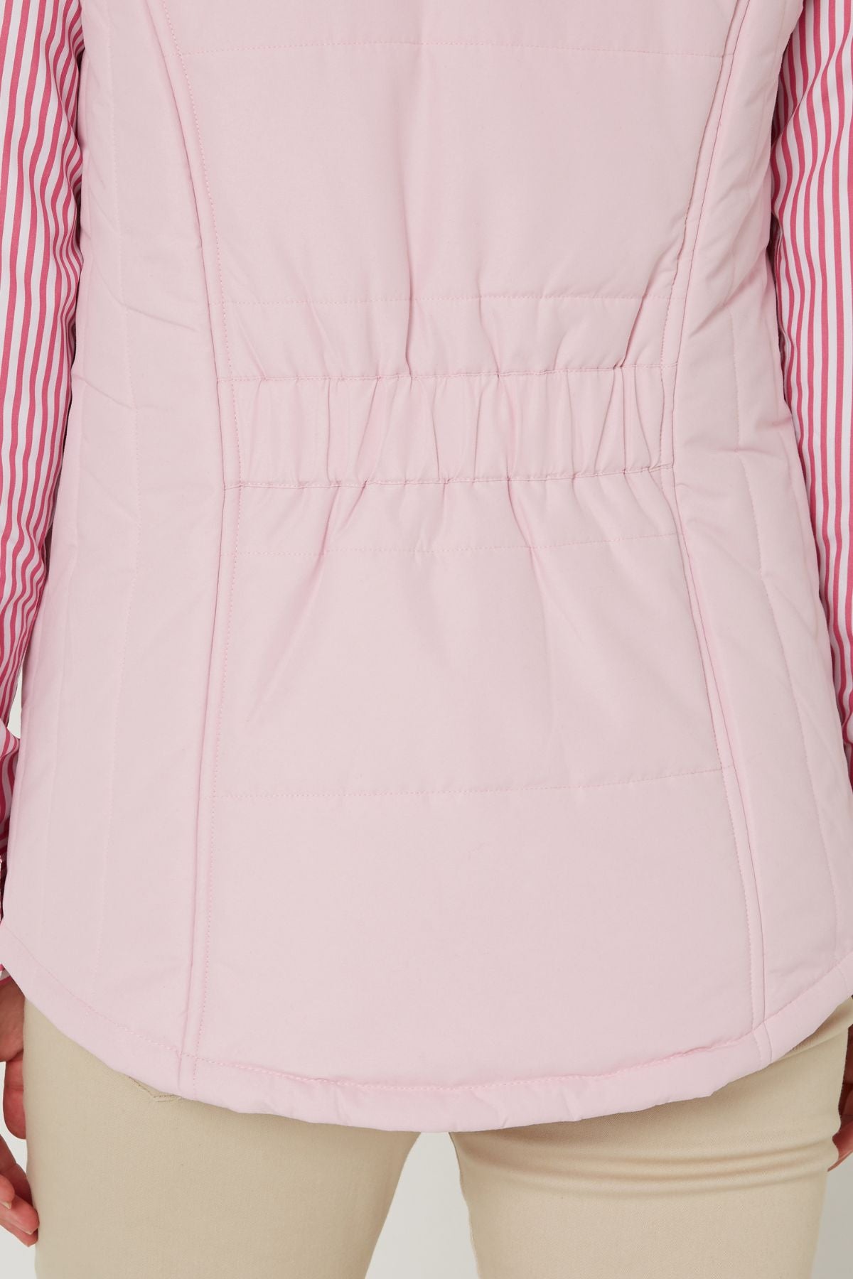 A close-up of the back of the Pilbara Ladies Vest in light pink with Polyfill padding and an elastic waistband, worn over a pink-and-white striped shirt and beige pants.