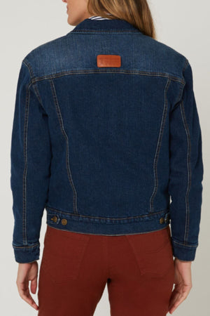 A person with long hair, seen from behind, wears a classic fit Pilbara Lauren Sherpa Lined Jacket in dark blue denim with a brown upper back patch, metal waistband buttons, and rust-colored pants.