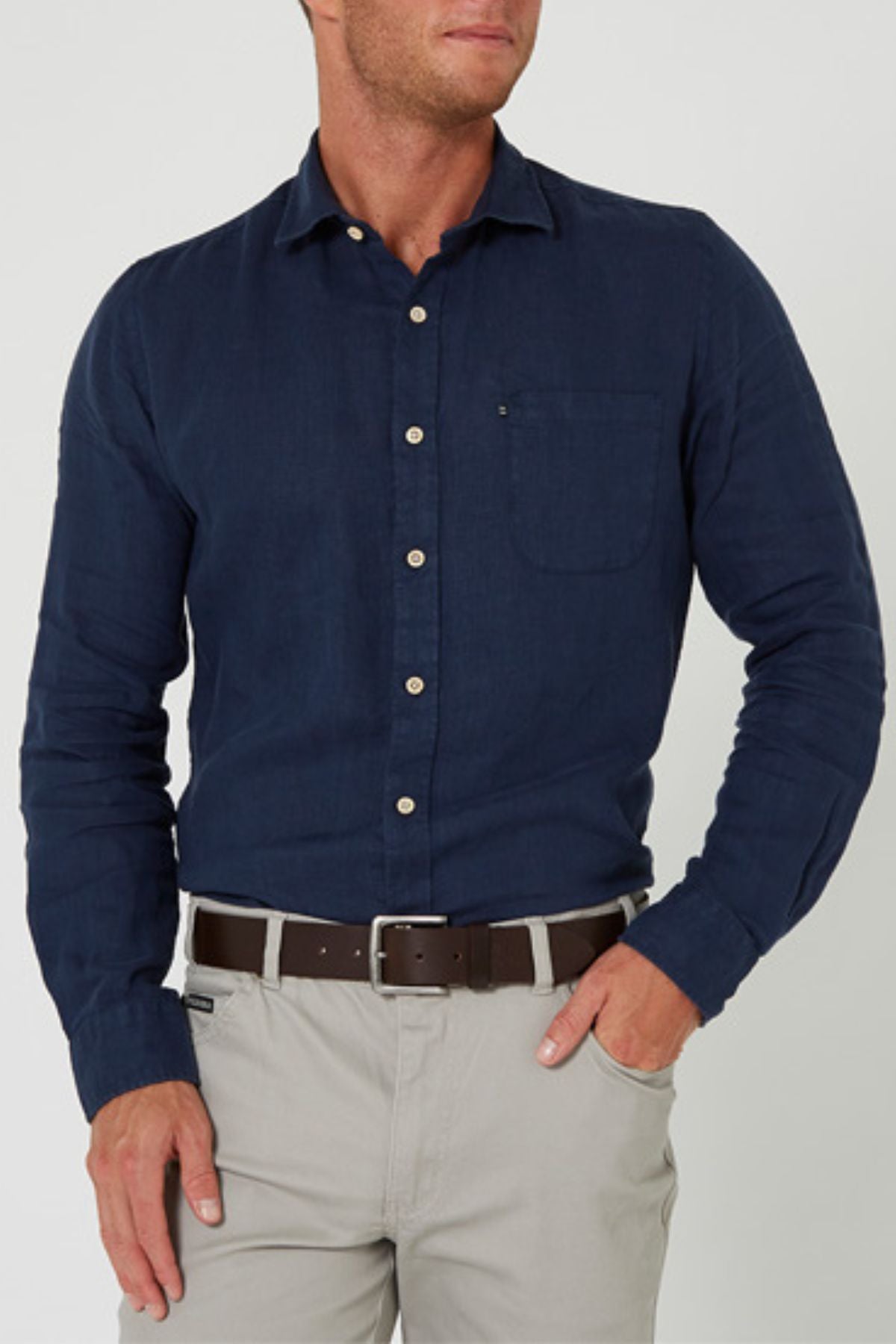 A man in a navy Pilbara Linen Shirt, made from 100% linen, and light gray pants stands with one hand in his pocket against a plain light background.