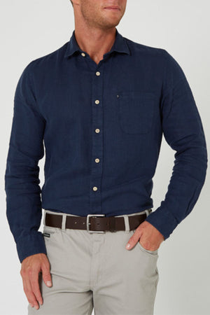 A man in a navy Pilbara Linen Shirt, made from 100% linen, and light gray pants stands with one hand in his pocket against a plain light background.