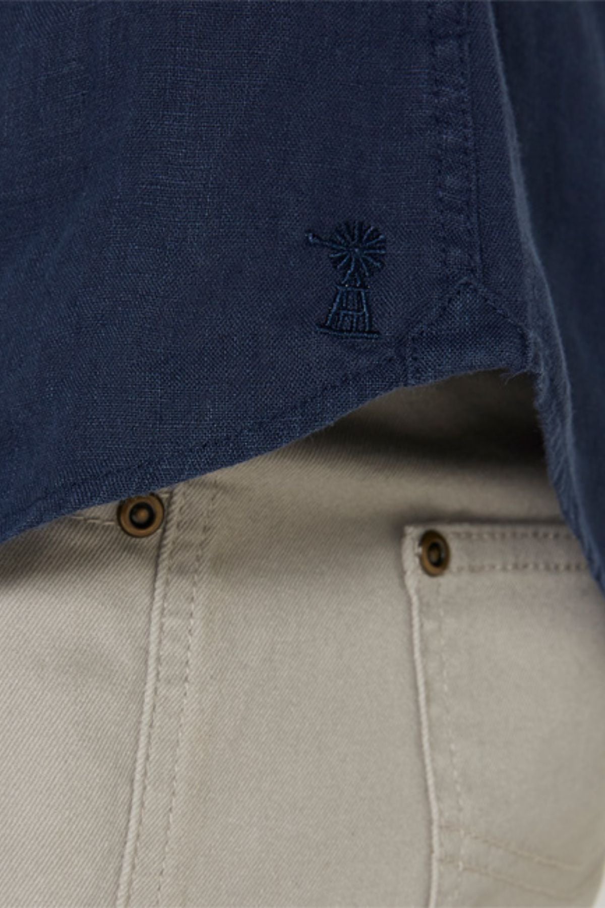 Close-up of the Pilbara Linen Shirt in navy blue, featuring a small windmill embroidery near the hem. Paired with light beige pants showcasing visible pockets and brown buttons.
