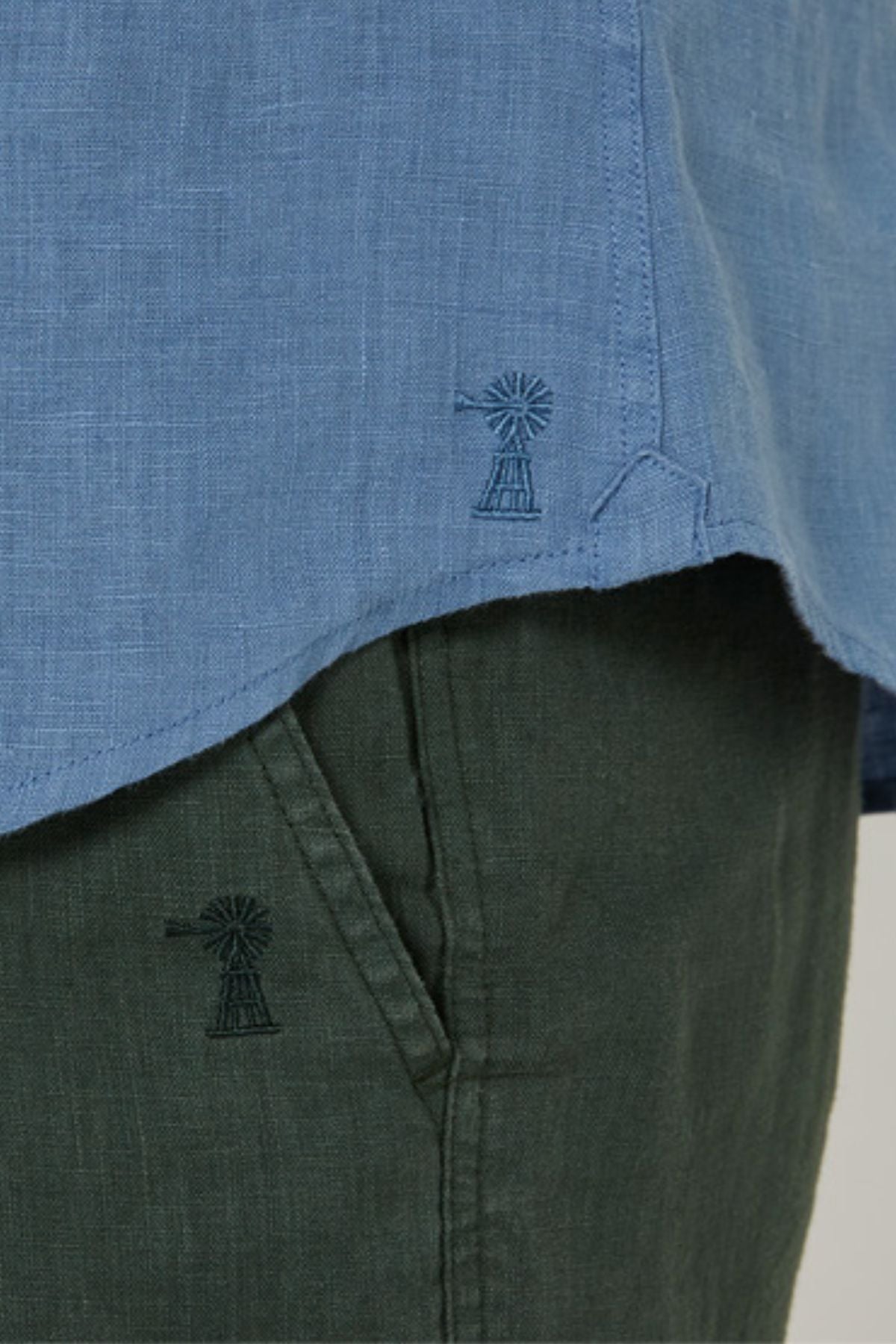 Close-up of a person wearing the Pilbara Linen Short Sleeve Shirt in light blue with dark green pants. Both the shirt hem and pants pocket feature matching windmill embroidery.