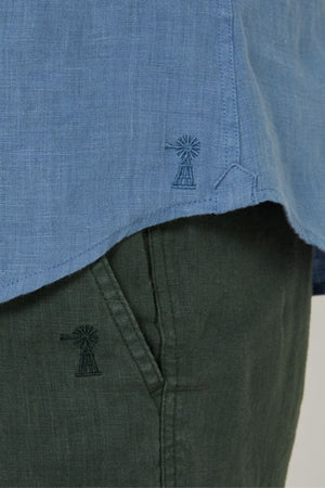 Close-up of a person wearing the Pilbara Linen Short Sleeve Shirt in light blue with dark green pants. Both the shirt hem and pants pocket feature matching windmill embroidery.