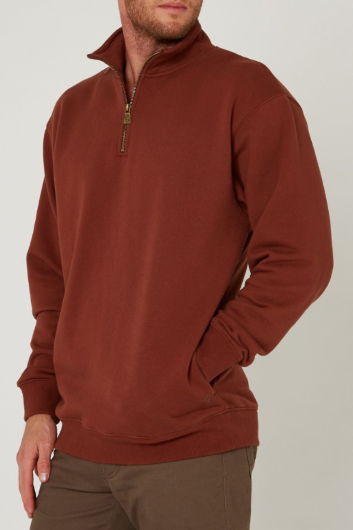 A man stands against a plain background with one hand in his pocket, wearing the Pilbara Classic Heavy Weight Fleece Pullover in rust and brown pants.