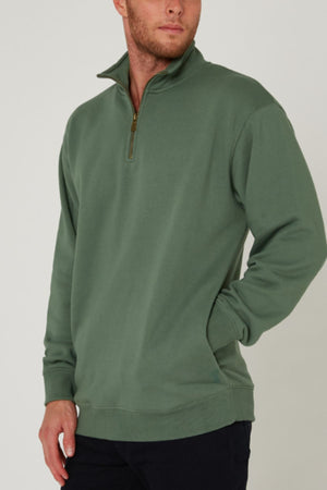 A man wearing a sage green Pilbara Classic Heavy Weight Fleece Pullover with a raised collar and black pants stands with one hand in his pocket, looking to the side against a plain light background.