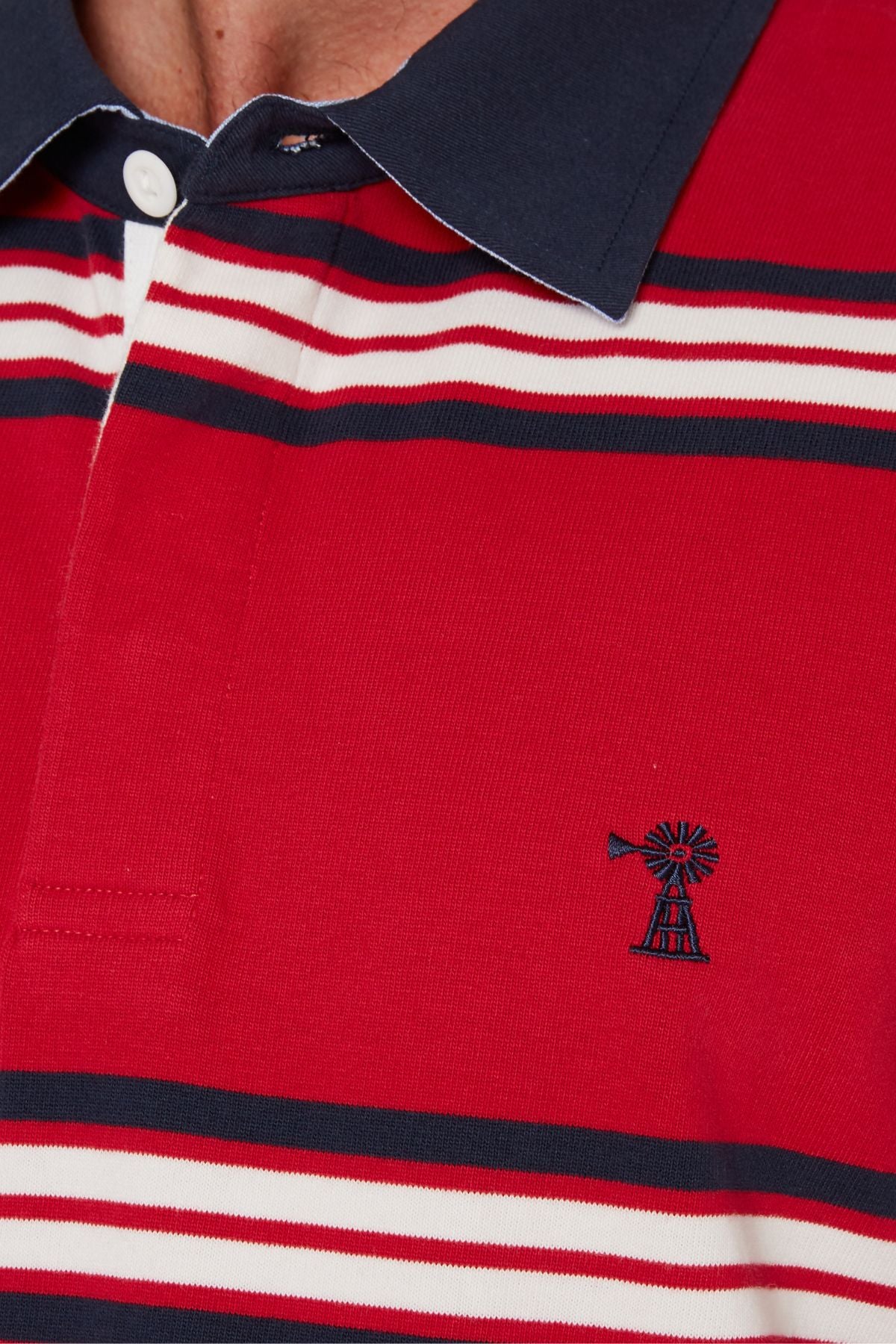 Close-up of the Pilbara Rugby Jersey, a red 100% cotton polo with white and navy horizontal stripes, a small windmill chest logo, and a navy collar with a white button, inspired by classic rugby designs.