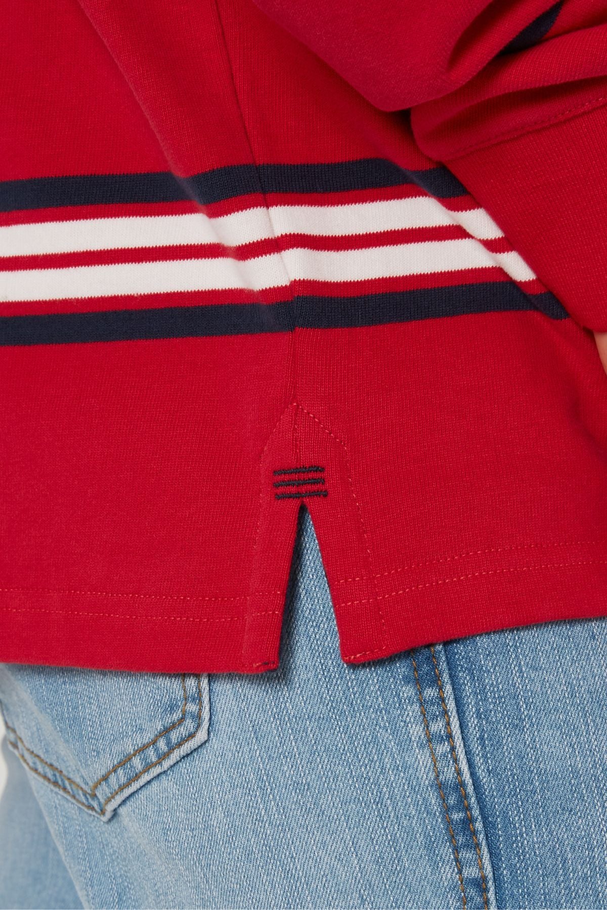 A close-up of a person in light blue jeans and a Pilbara Rugby Jersey, made from 100% cotton, in red with white and navy horizontal stripes, featuring a small side slit with extra navy blue stitching.