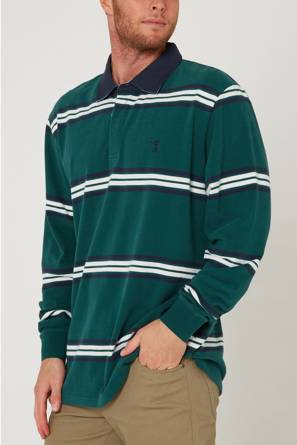 A man wearing the Pilbara Rugby Jersey—a green long-sleeve shirt with white and navy stripes made of 100% cotton—and khaki pants stands with one hand in his pocket against a plain background.
