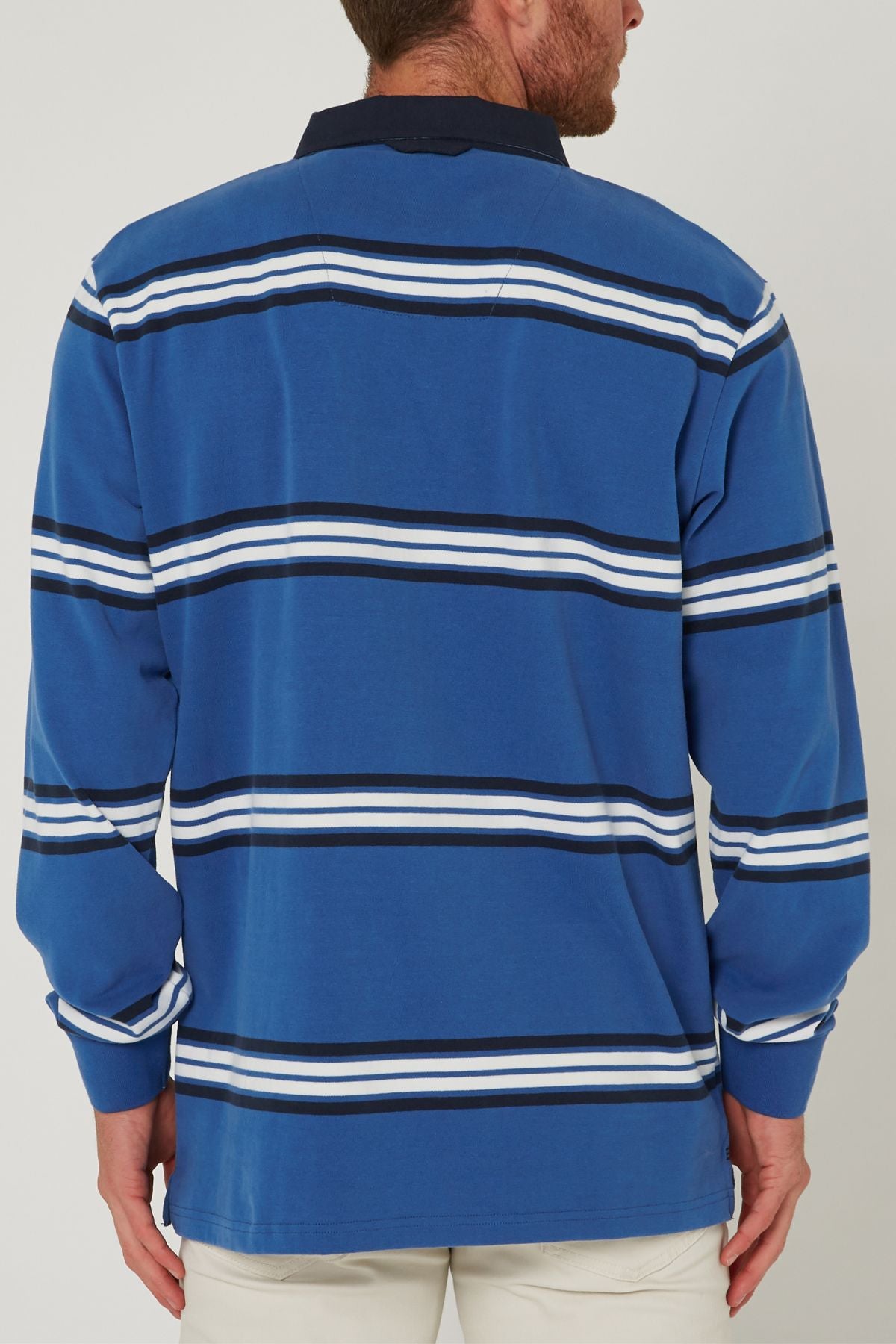 A man is seen from behind wearing a Pilbara Rugby Jersey in blue with white and black horizontal stripes, a dark collar, and white pants.