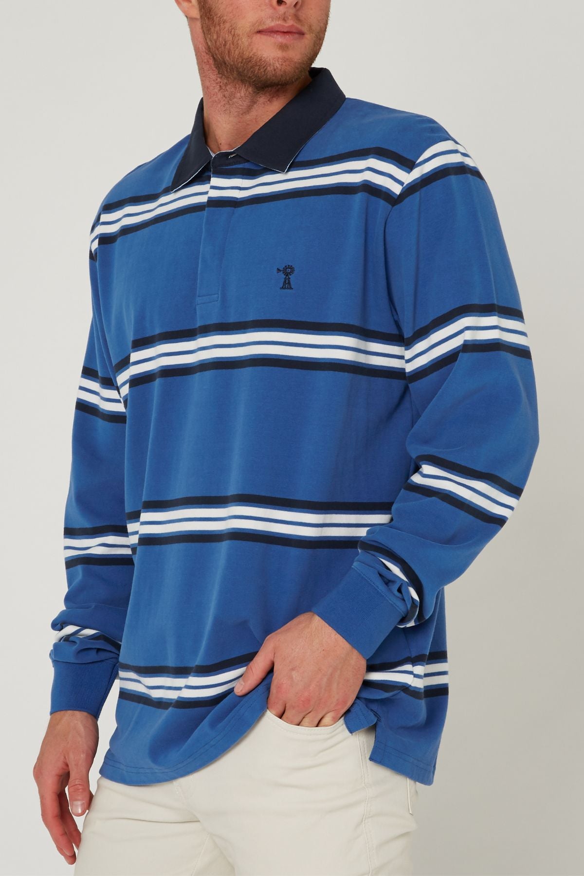 A person wears the Pilbara Rugby Jersey—blue with white and navy stripes, featuring a small left-chest logo—paired with light-colored pants. Their face is partially out of frame.