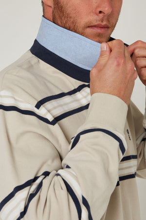 A person adjusts the blue denim collar of a beige Pilbara Rugby Jersey with navy and white horizontal stripes. Only the lower half of their face and upper torso are visible.