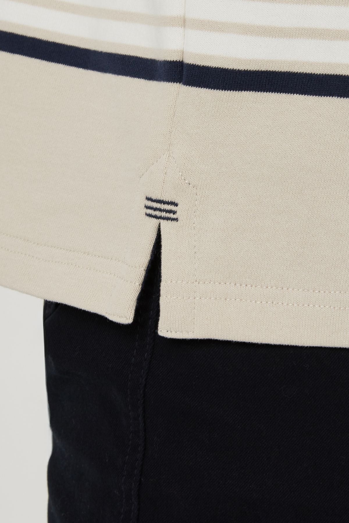 Close-up of the hem of the Pilbara Rugby Jersey, a beige shirt with dark blue stripes, classic rugby style, featuring a small side slit and decorative stitching, worn over dark pants.