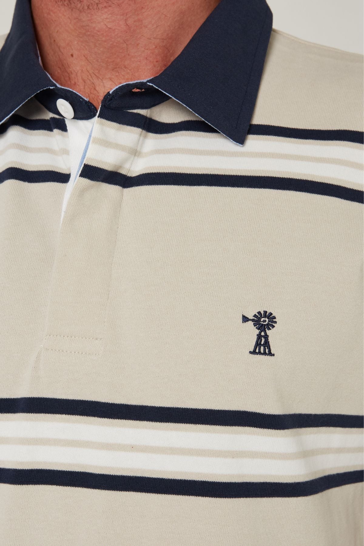 Close-up of a person wearing the Pilbara Rugby Jersey, featuring beige, navy, and white stripes, a navy collar, and an embroidered windmill logo on the chest—classic style with subtle rugby inspiration.