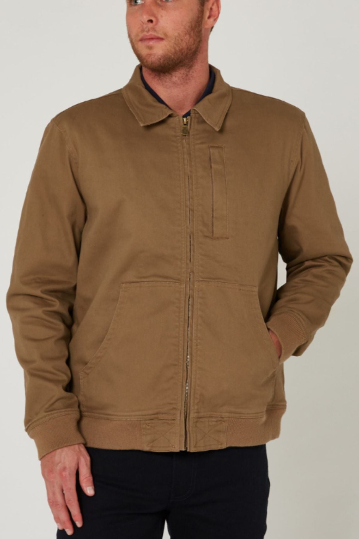 A man stands against a plain light background wearing the Pilbara Sherpa Bomber Jacket, a tan cotton stretch twill piece with front pockets and a collar, one hand in his pocket.