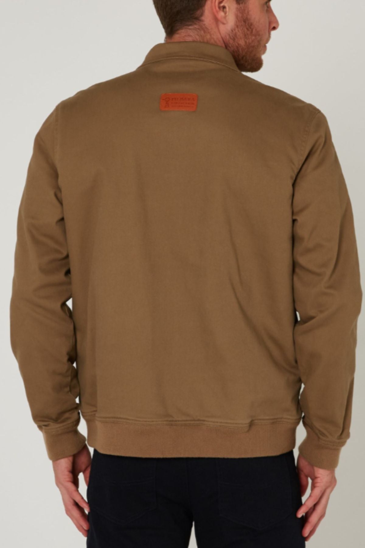 A man is seen from behind wearing a tan Pilbara Sherpa Bomber Jacket with a small rectangular patch on the upper back and dark pants, the sherpa-lined bomber standing out against a plain, light-colored background.
