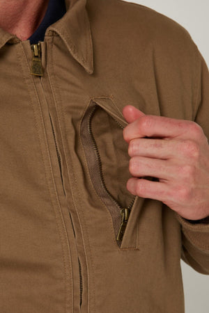 A person unzipping the chest pocket of the Pilbara Sherpa Bomber Jacket, a tan men’s cotton stretch twill jacket with a visible collar and brass-colored zippers, blending classic style and comfort.