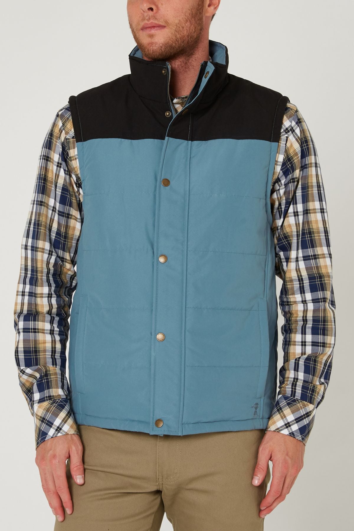 A man stands against a plain light background wearing the Pilbara Two Tone Vest over a plaid button-up shirt and khaki pants.