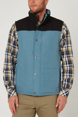 A man stands against a plain light background wearing the Pilbara Two Tone Vest over a plaid button-up shirt and khaki pants.