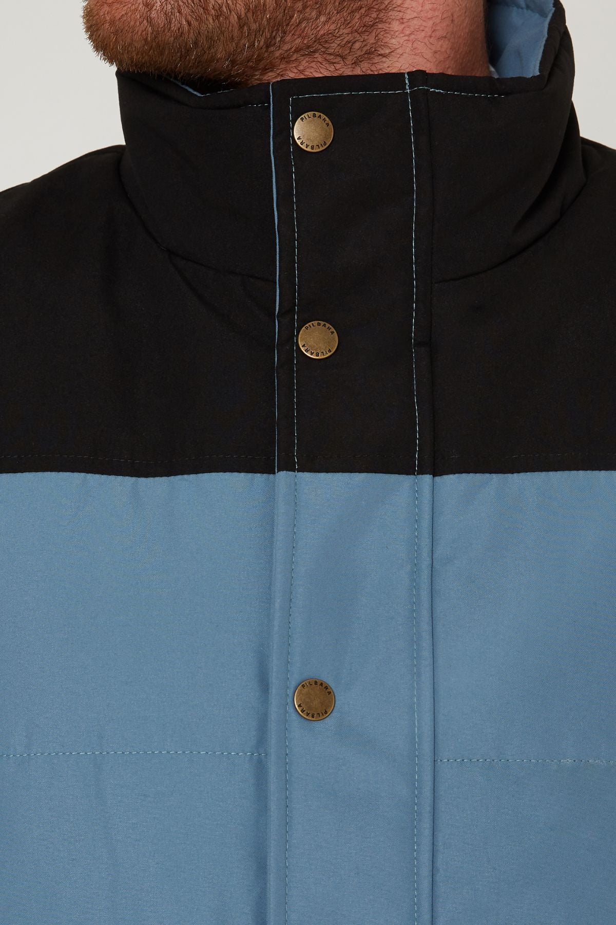 Close-up of a person wearing the Pilbara Two Tone Vest, featuring polyfill padding, a black top section, blue lower section, and metal snap buttons. Only the person's lower face and neck are shown.