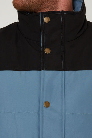 Close-up of a person wearing the Pilbara Two Tone Vest, featuring polyfill padding, a black top section, blue lower section, and metal snap buttons. Only the person's lower face and neck are shown.